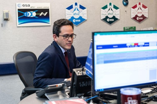 A person sitting at a desk on a computer wearing a suit with red, green, and blue mission patches in the background.